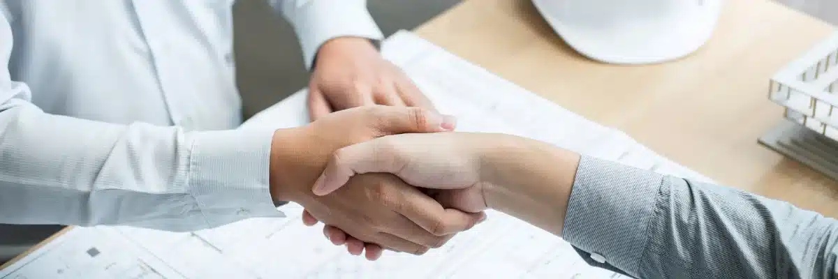 Un artisan et un partenaire se serrent la main au-dessus de plans, deux professionnels qui se recommandent